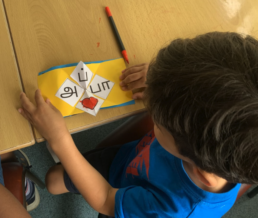 Child arranging Tamil letter pieces on a colourful paper craft at a classroom table